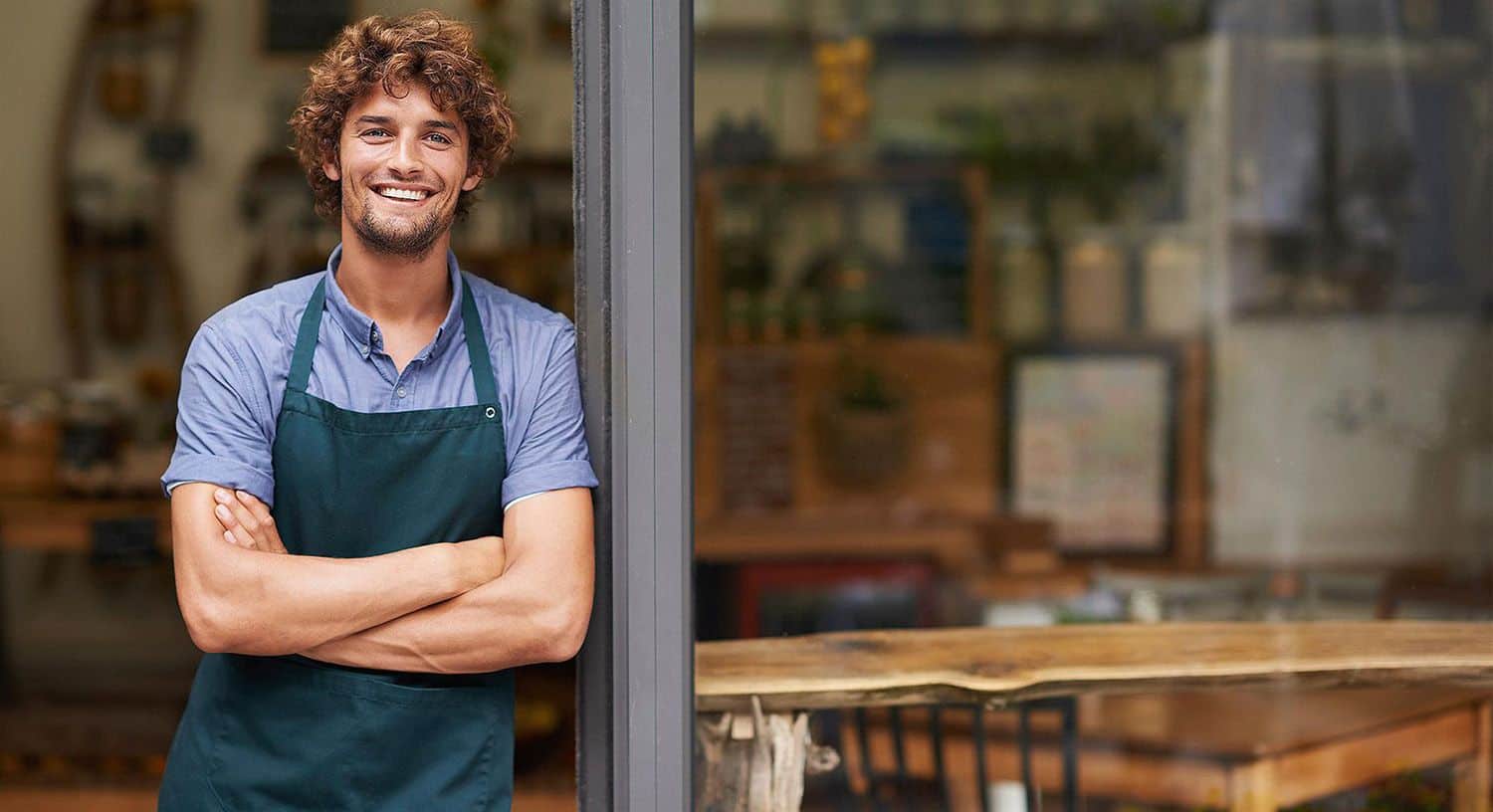 Confident, curly-haired man in blue shirt and green apron welcomes guests at cozy café—ideal for agency branding or websites.