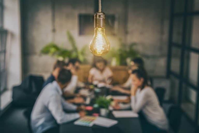 Close-up of a glowing light bulb overhead, with a blurred team meeting in the background, symbolizing innovation and creative strategy.