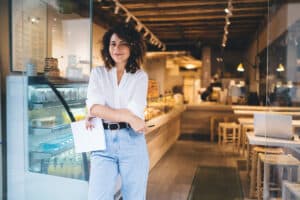 A woman with curly hair and a white shirt stands at a modern café entrance, smiling confidently as she holds papers for web marketing.
